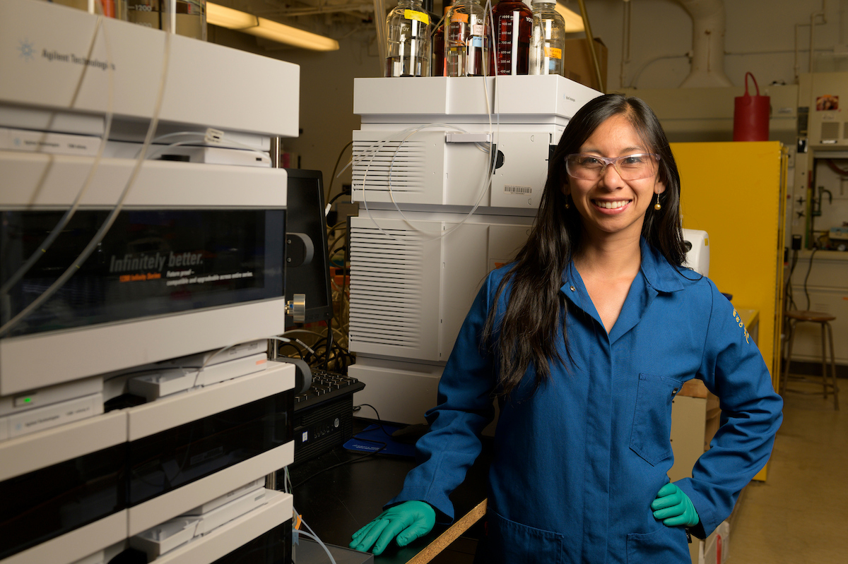 Smiling woman scientist in blue lab coat and gloves beside large chemistry lab instruments