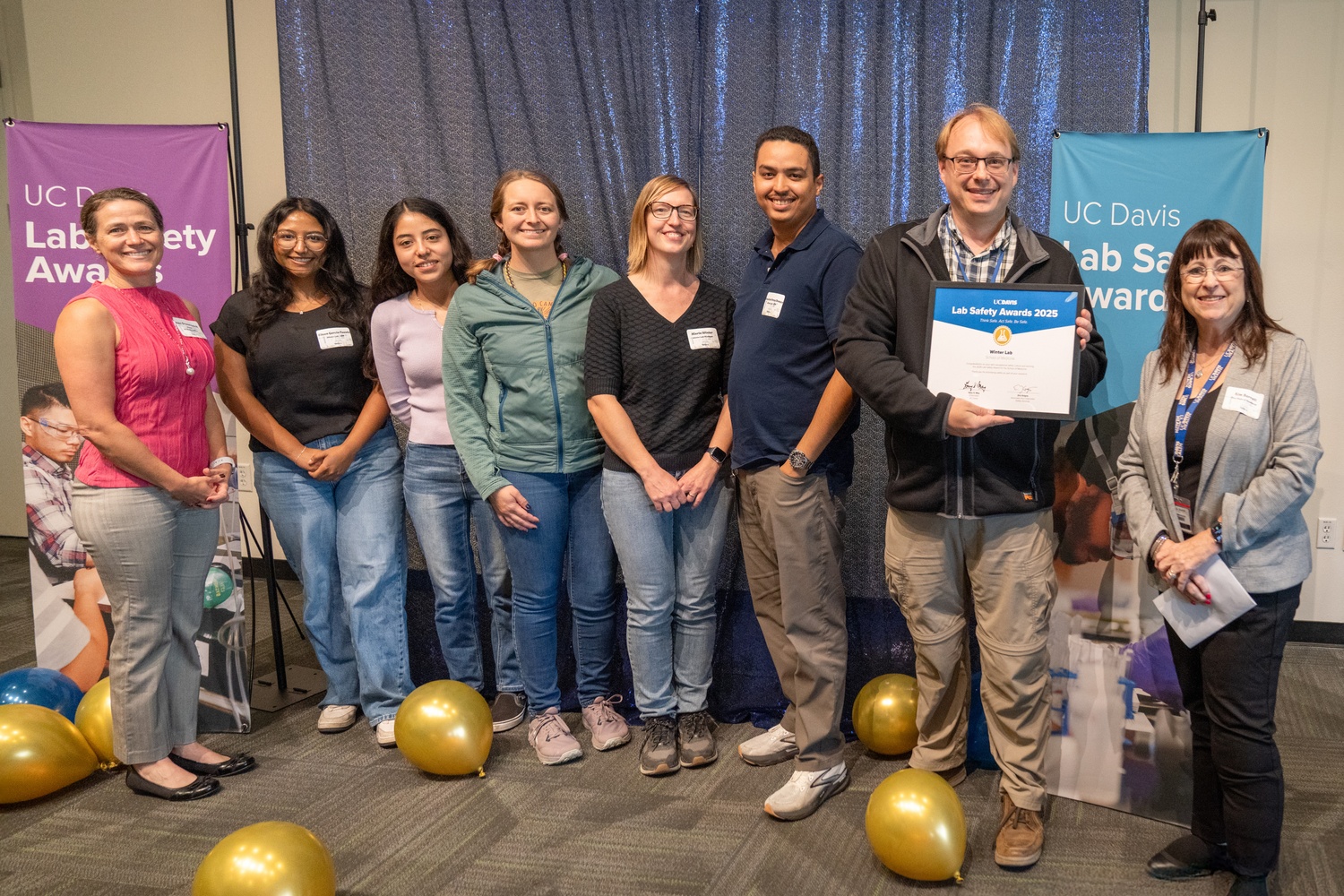 Members of the Winter Lab smile with their Lab Safety Award Certificate against a blue backdrop and award banners.