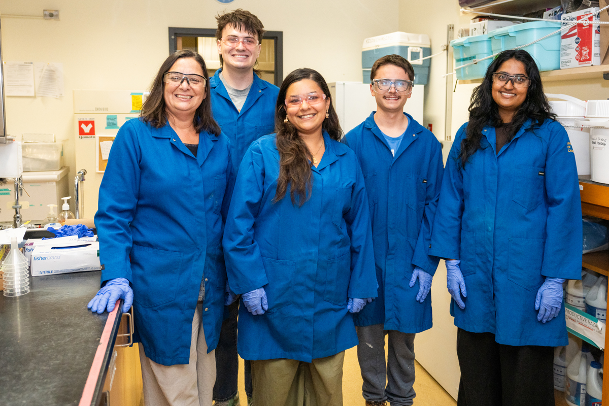 Five lab members in blue University of California lab coats and gloves smiling in a Plant Sciences lab