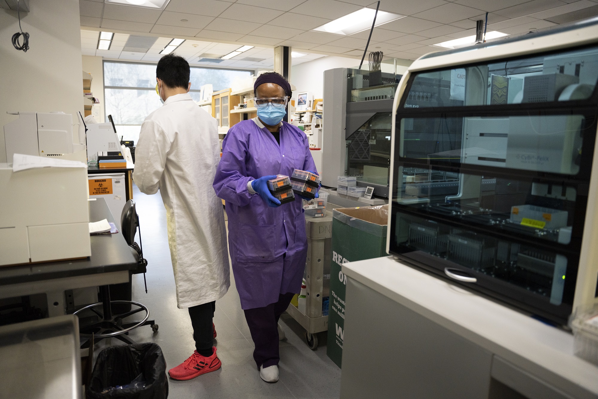 lab workers passing each other between lab benches. Both are wearing correct PPE. 