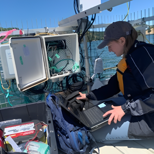 Researcher uses a laptop and is surrounded by other equipment while on a boat in a reservoir.