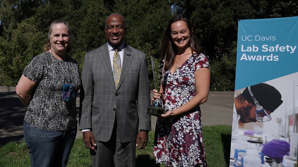 Dr. Rachael Bay (right) holding Lab Safety Awards trophy and pictured with Lab Manager and Departmental Safety Coordinator, Brenda Cameron (left), and Chancellor Gary May.