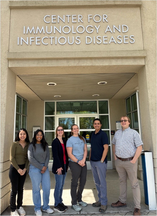 Dr. Sebastian Winter (right) and five other people stand together and smile outside a building with the sign "Center for Immunology and Infectious Diseases"