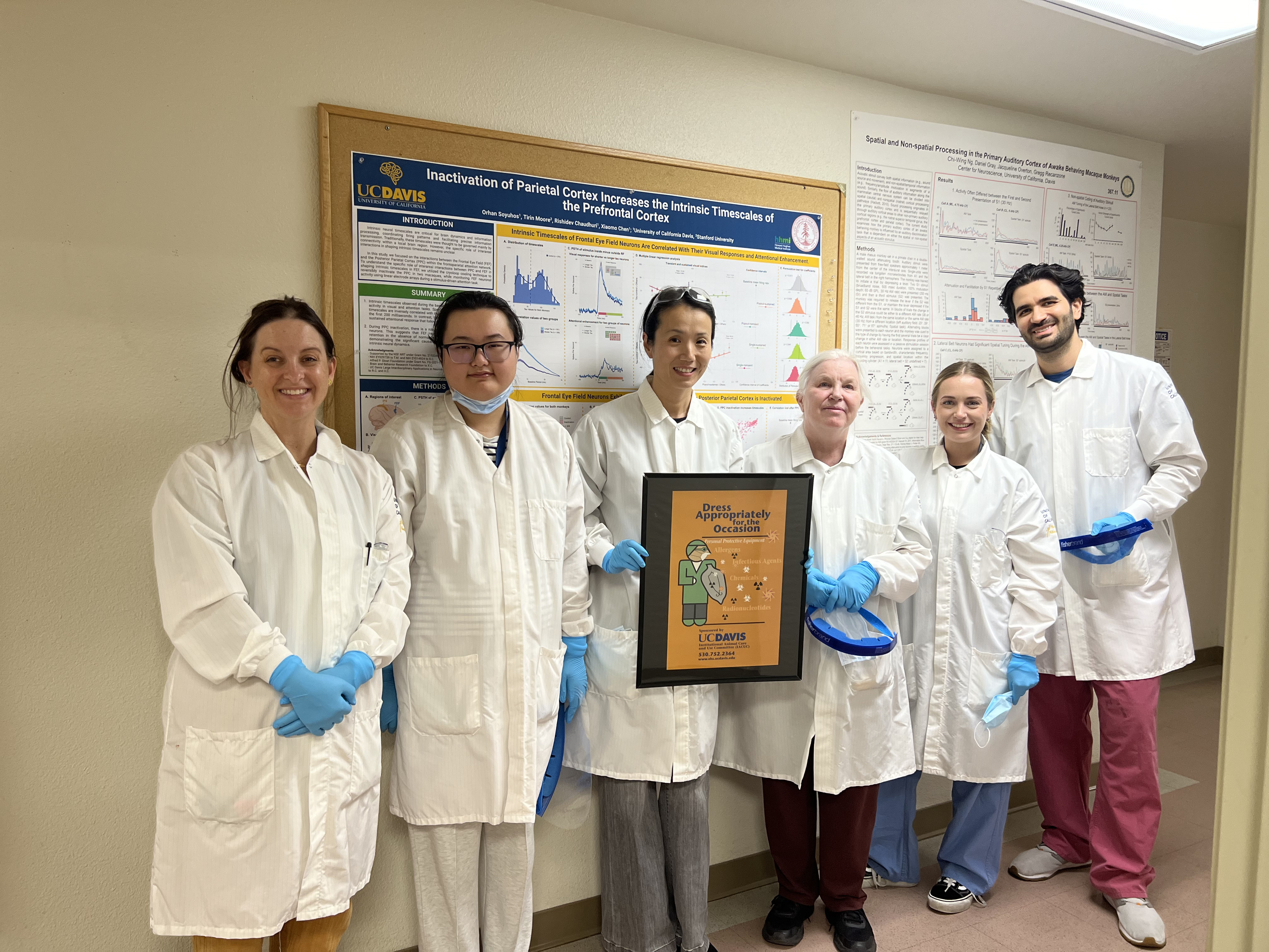 Dr. Chen and her lab group in white lab coats and holding a sign that reads "Dress appropriately for the occasion."