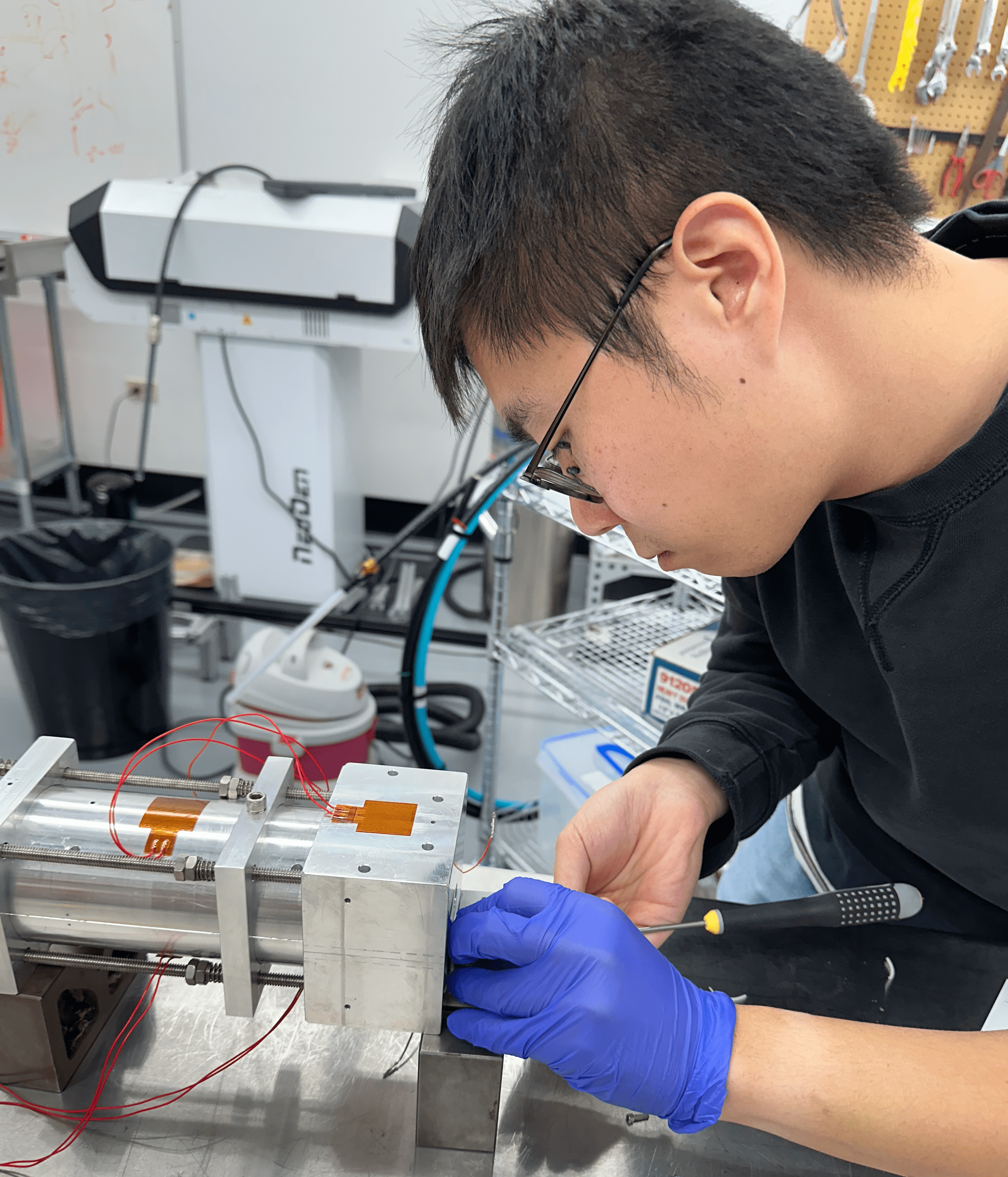 A man wearing glasses and a blue glove uses a screwdriver on a piece of metal machinery.