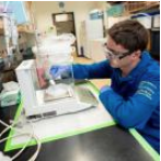Student in blue lab coat and goggles working in a lab