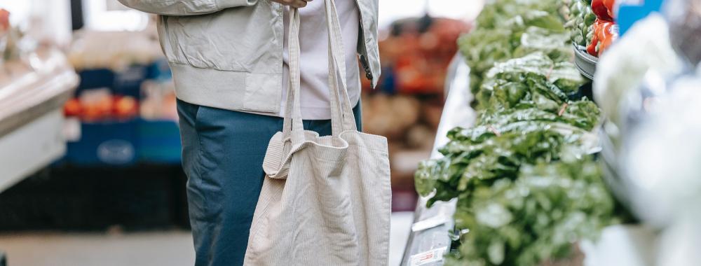 person wearing a gray coat holding a reusable shopping bag standing next to green vegetables