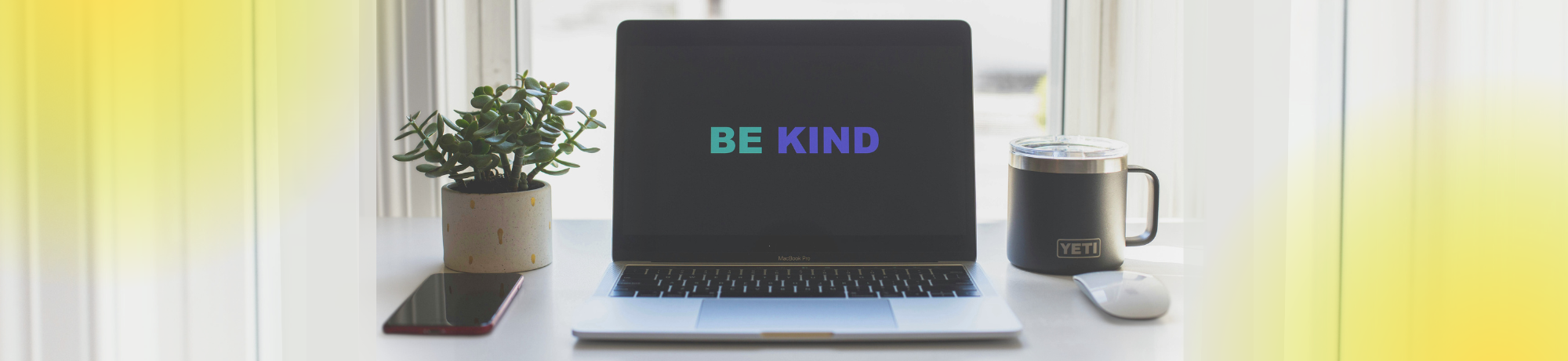 Laptop screen with be kind written across screen on tabletop with plant, mug, mouse and phone