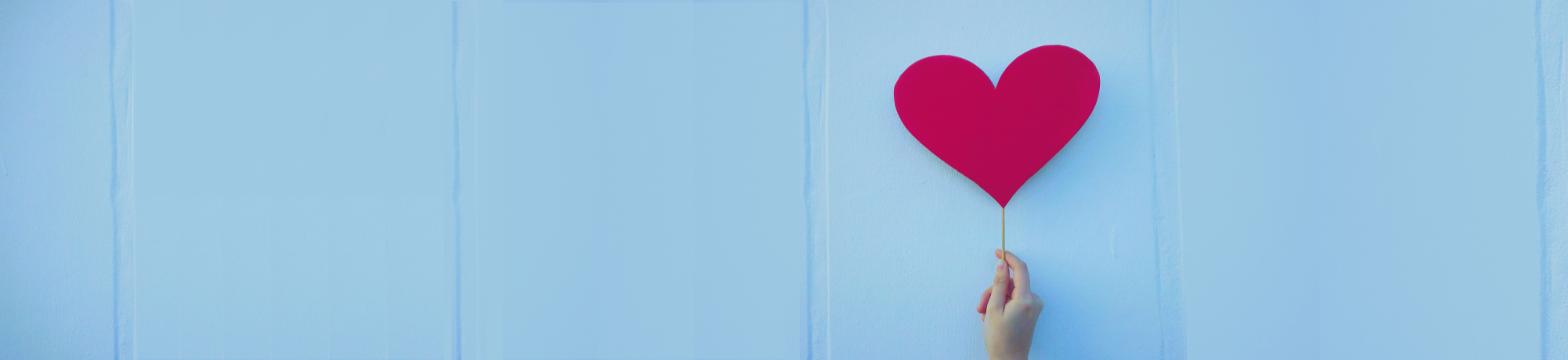 Person's hand holding up a red paper heart on a stick against an exterior light blue wall