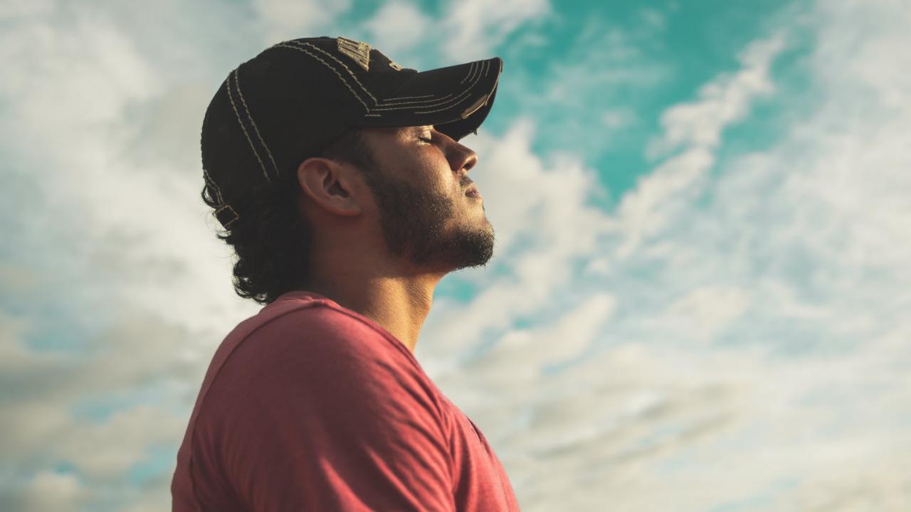 Person with eyes closed, wearing a baseball cap standing and facing toward the sun. 