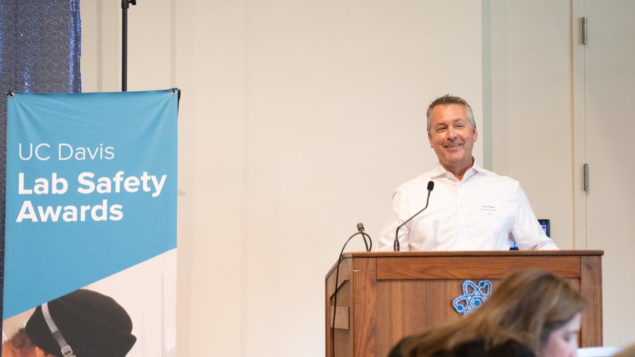 Associate Vice Chancellor Eric Kvigne stands at a lecturn and speaks. Beside him is a blue banner that reads "UC Davis Lab Safety Awards."