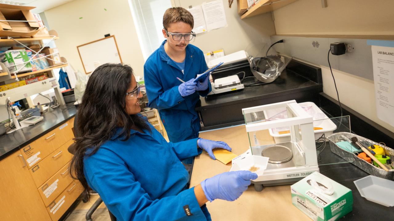 Two UC Davis students in blue lab coats weighing and recording a field sample