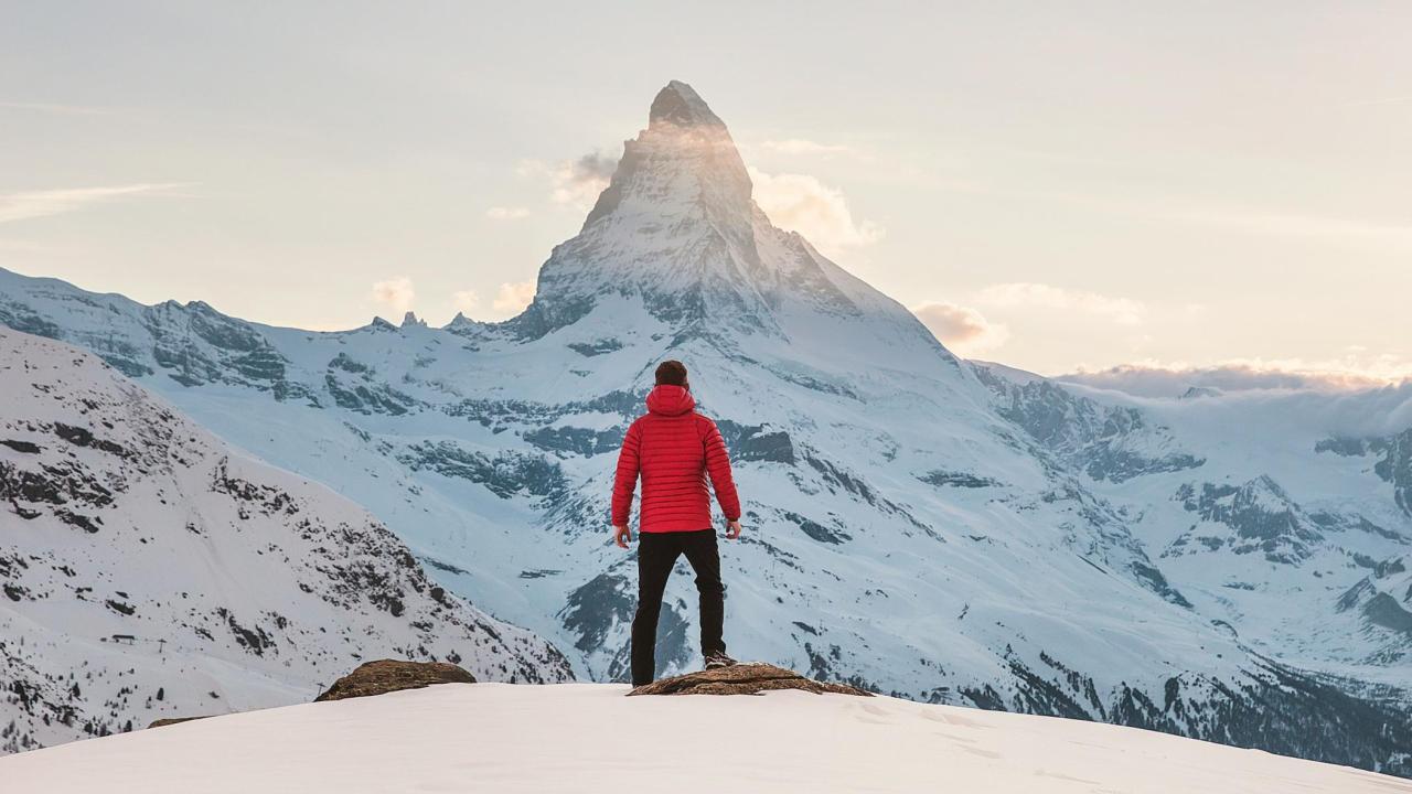 Image of a person with their back to the camera facing a snowy mountain top wearing a red coat