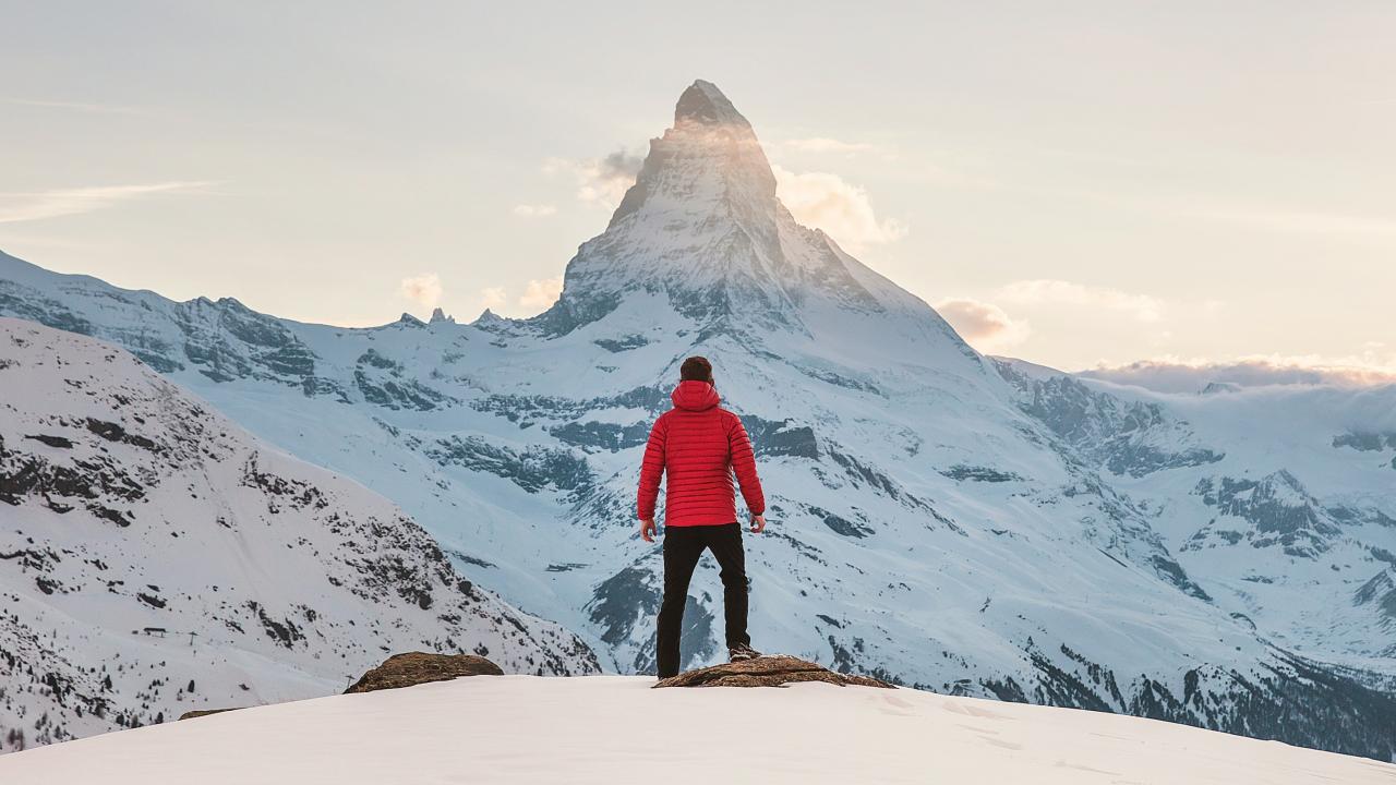 Image of a person with their back to the camera facing a snowy mountain top wearing a red coat