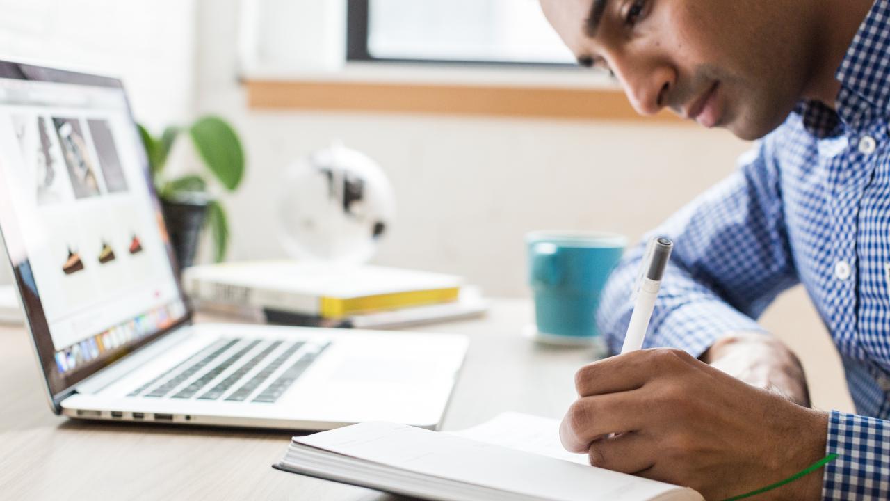 person writing notes in a journal in front of laptop