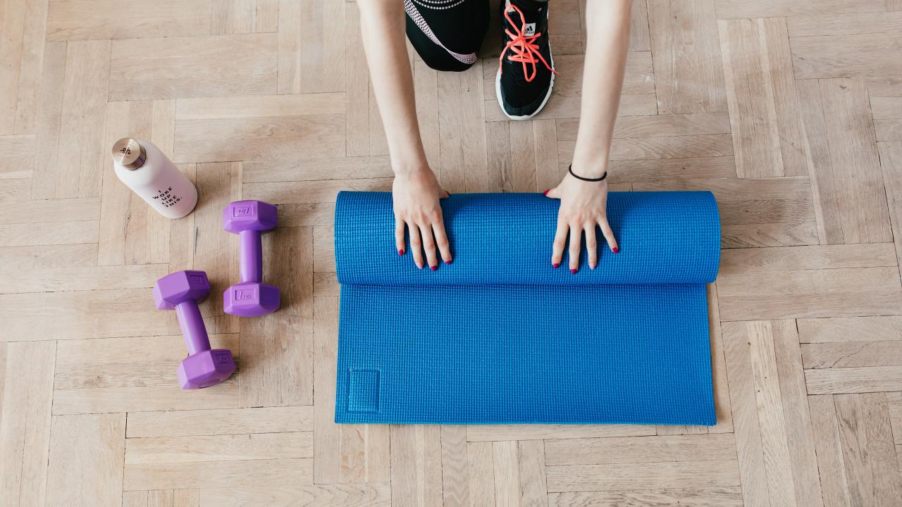 person rolling out yoga mat with a water bottle and purple dumbbells on the floor to their right
