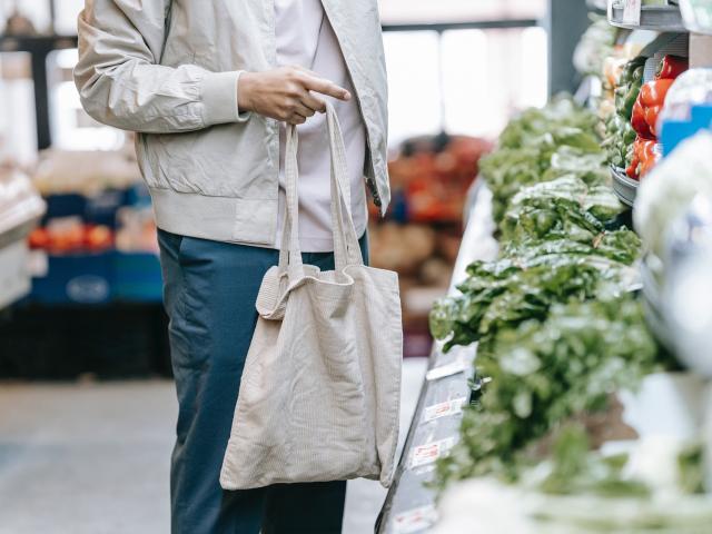 person wearing a gray coat holding a reusable shopping bag standing next to green vegetables