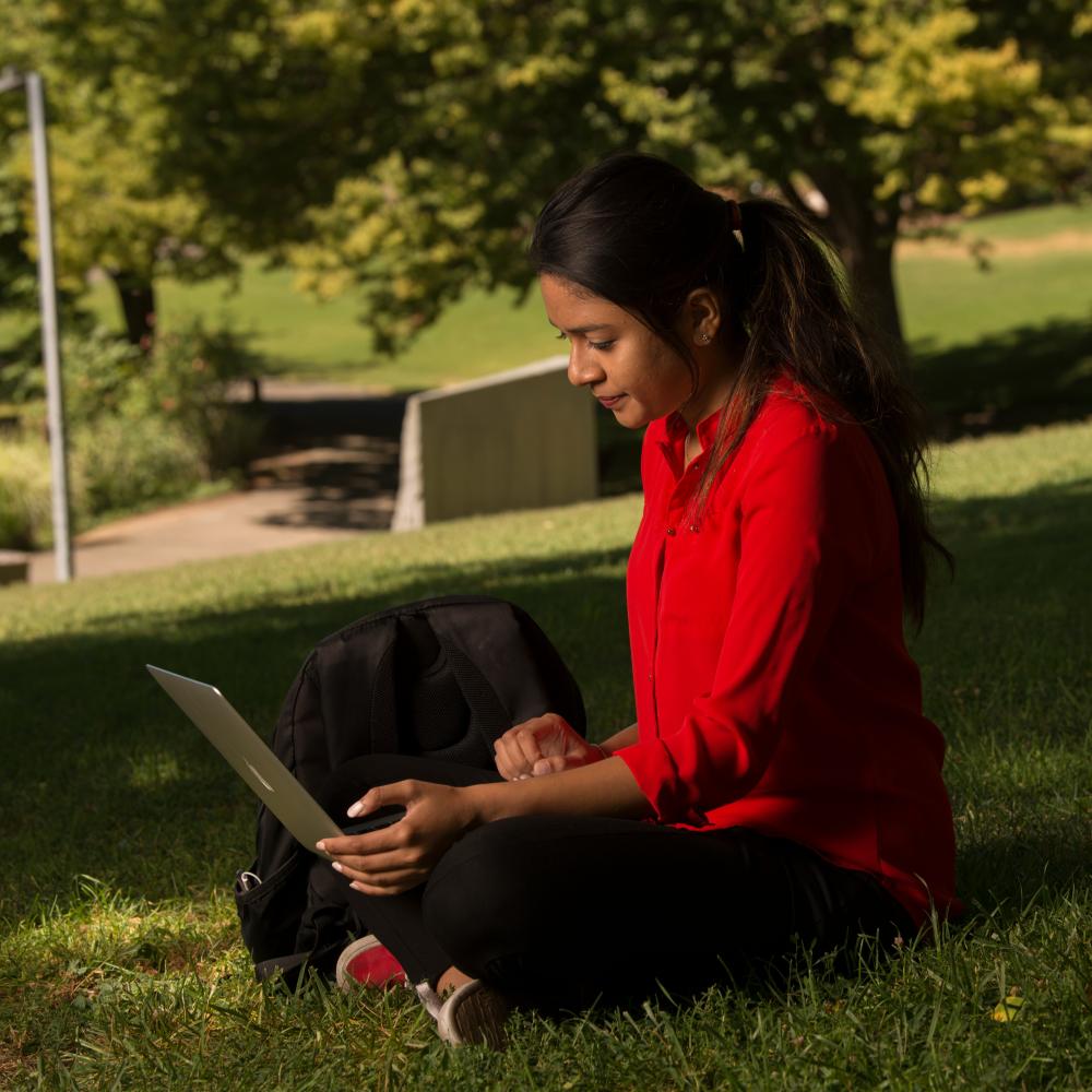 A woman in a red shirt sits on the lawn while looking at her laptop