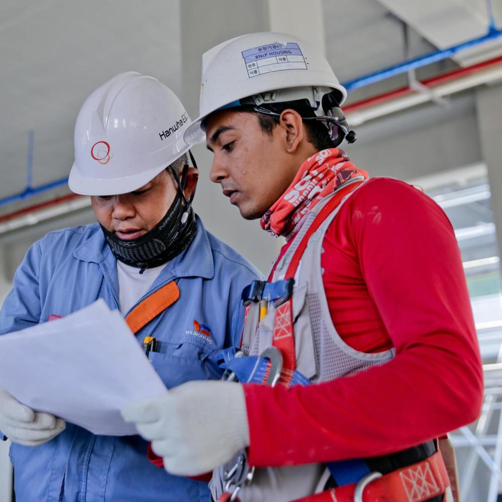 Two men in hard hats look at a piece of paper. In the background, construction is underway.