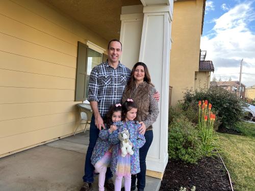 A dark-haired man and brunette woman stand behind their two young daughters.