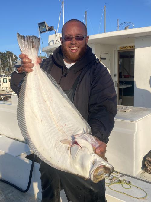 Nate Arceneaux on a boat in San Francisco Bay holding a CA halibut fish he caught. The fish is flat, large and white. 