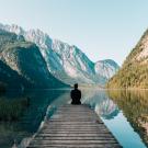 person sitting on a lake dock looking toward mountains