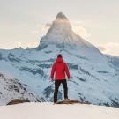 Image of a person with their back to the camera facing a snowy mountain top wearing a red coat