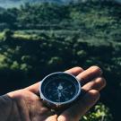 close up of a person's hand holding a compass against a lush green landscape in the background